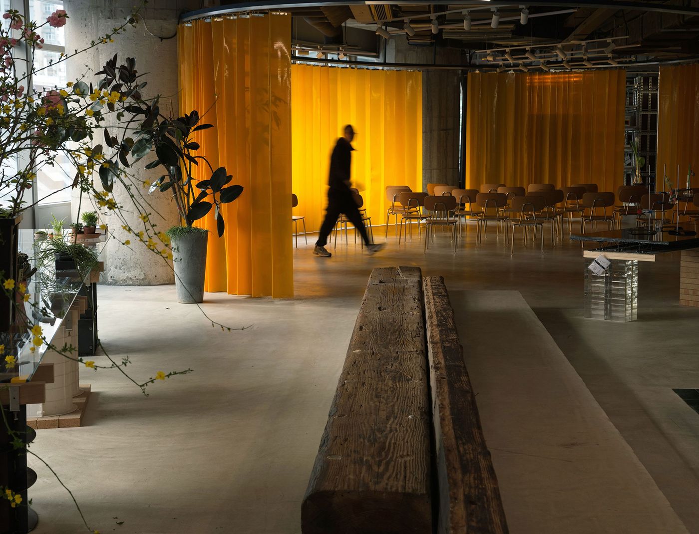 A dynamic industrial-chic interior in the 889 GLO Art Space, Shanghai, features a vibrant yellow curtain acting as a flexible partition, casting a warm glow. A blurred figure walks past a raw wooden bench and sparse seating, highlighting the interplay of robust materials—concrete, wood, and metal—with vivid color, creating a versatile, evocative atmosphere.