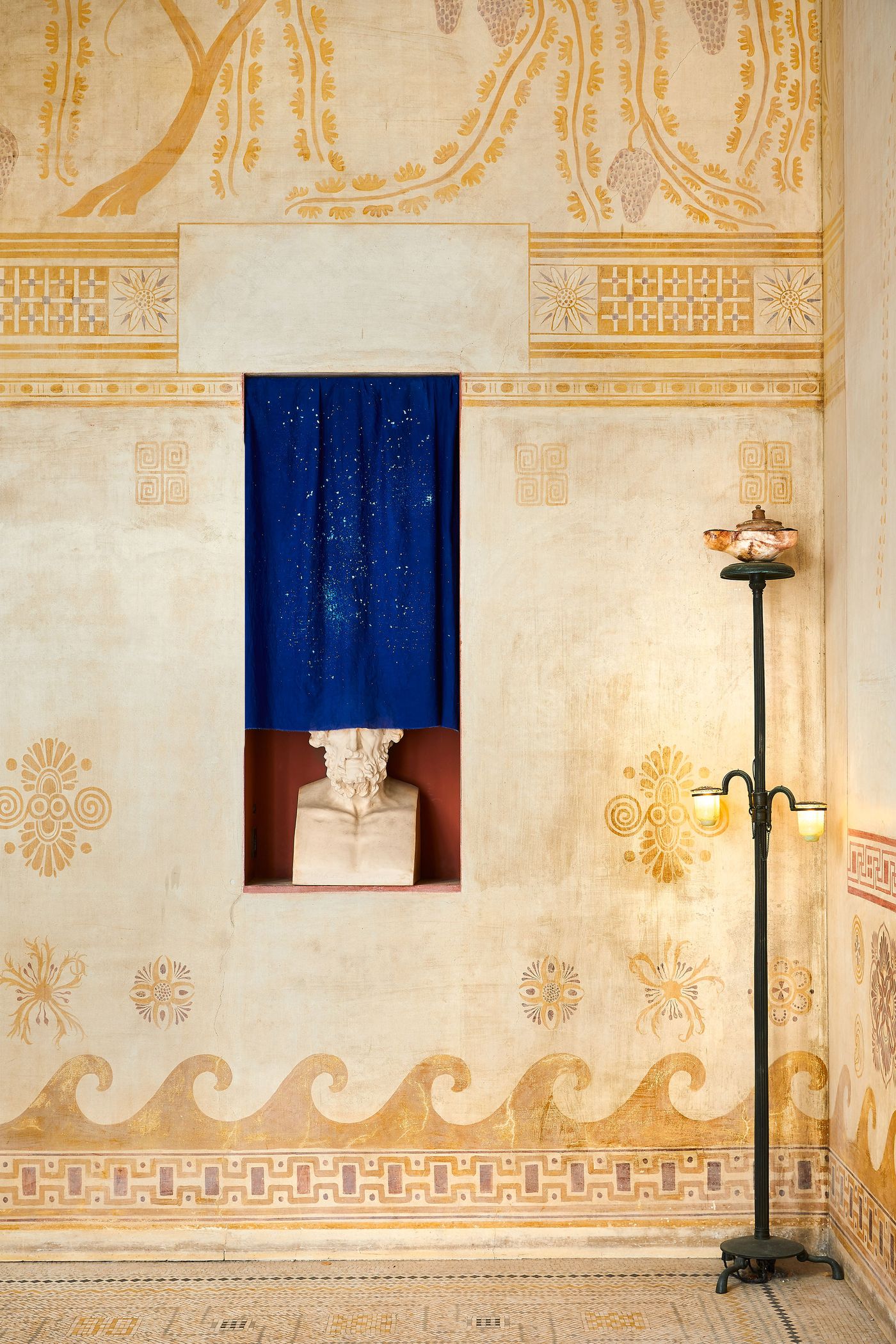 A section of a room at Villa Kerylos featuring a decorative niche with a white marble bust, half-obscured by a vibrant blue curtain speckled with white. The surrounding walls are a canvas of warm tones, adorned with biomorphic and geometric murals in a neo-vernacular style. A section of a room at Villa Kerylos featuring a decorative niche with a white marble bust, half-obscured by a vibrant blue curtain speckled with white. The surrounding walls are a canvas of warm tones, adorned with biomorphic and geometric murals in a neo-vernacular style.