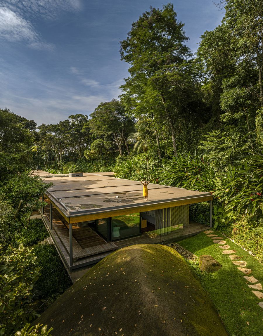An aerial view highlights the sleek, low-profile architecture, designed to immerse itself in the dense Brazilian rainforest. The minimalist structure with its expansive flat roof and floor-to-ceiling glass walls is surrounded by lush green foliage and natural rock formations, creating a striking biophilic sanctuary. A figure stands on the rooftop, embracing the serene natural vista. An aerial view highlights the sleek, low-profile architecture, designed to immerse itself in the dense Brazilian rainforest. The minimalist structure with its expansive flat roof and floor-to-ceiling glass walls is surrounded by lush green foliage and natural rock formations, creating a striking biophilic sanctuary. A figure stands on the rooftop, embracing the serene natural vista.