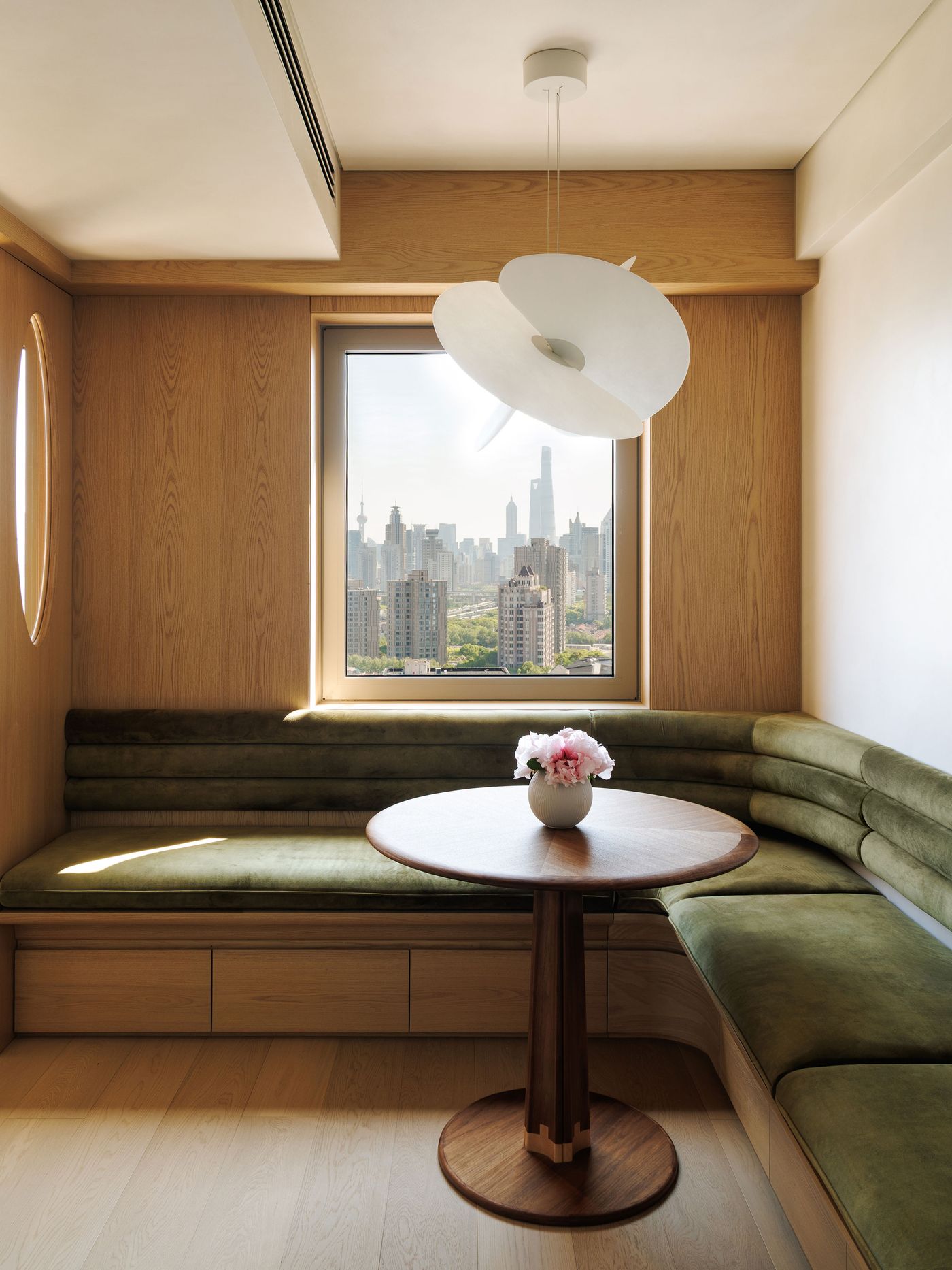 An intimate dining area bathed in natural light from a large window overlooking the Shanghai skyline. The space features a custom-built, U-shaped banquette upholstered in rich olive green velvet, complementing the light wood paneling. A sculptural white pendant light hovers over a round wooden table, adorned with pink peonies, creating a serene and sophisticated urban retreat.