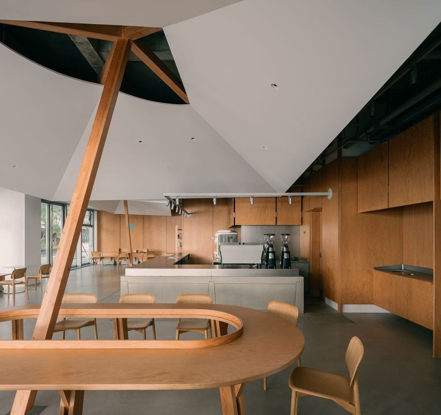 A panoramic view of the café unveils its spatial rhythm: concrete floors, soft-hued woodwork, and geometric ceiling cuts converge around a sculptural wooden table with an open oval core.
