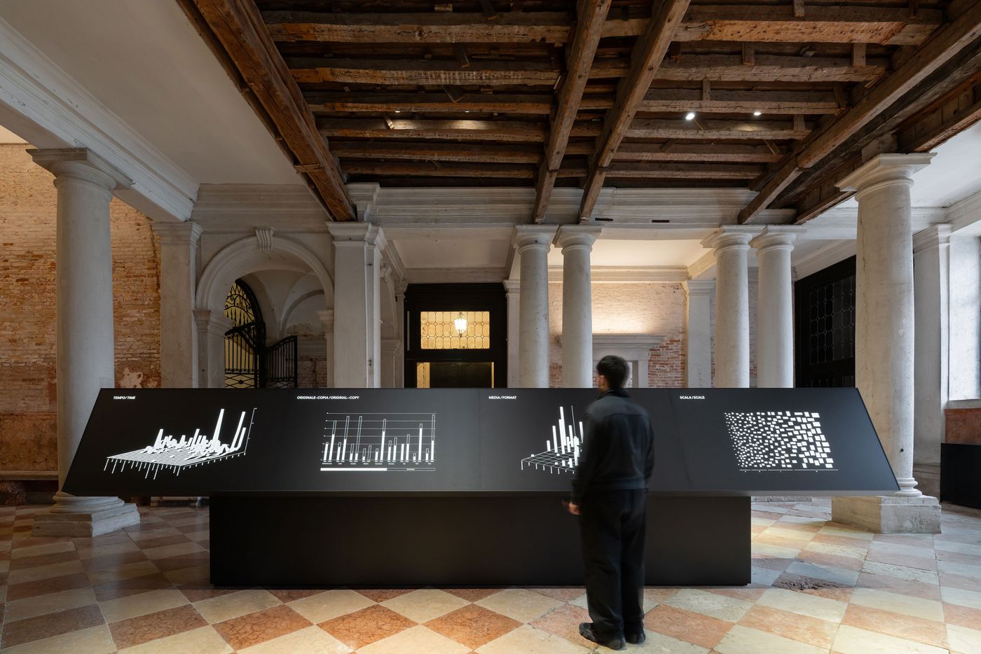 A visitor stands before a sleek exhibit displaying three data visualizations in a spacious, historic gallery with wooden beams. A visitor stands before a sleek exhibit displaying three data visualizations in a spacious, historic gallery with wooden beams.
