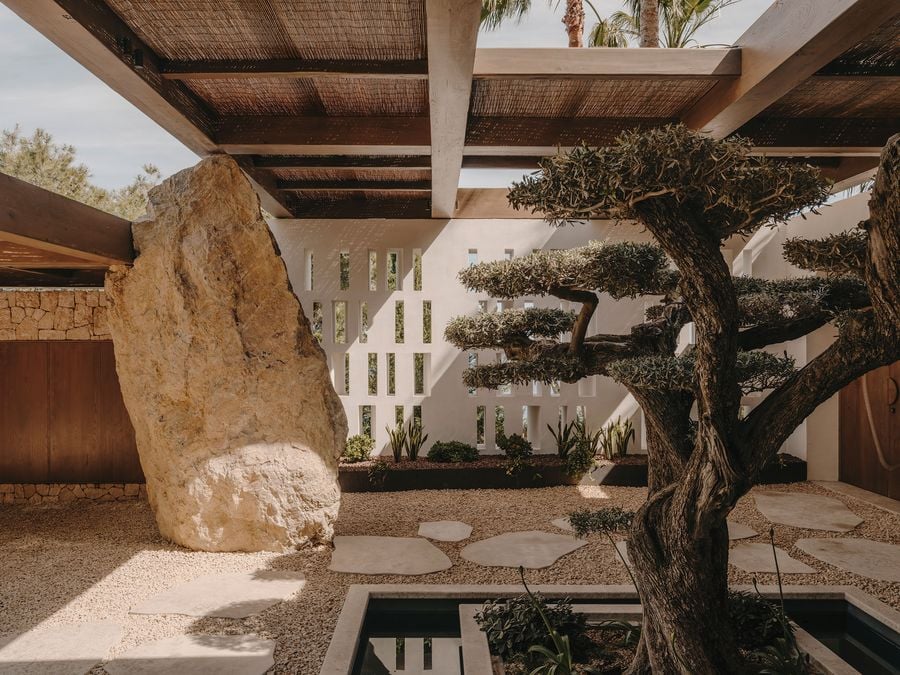 A serene outdoor space featuring a large rock, sculpted tree, and modern architecture with white walls and stone pathways.