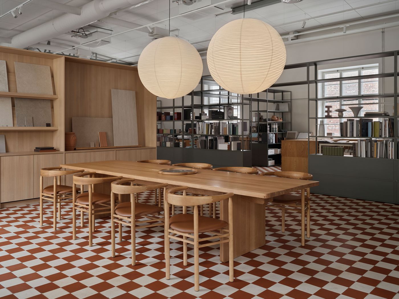 A large, natural wood dining table with accompanying chairs sits prominently on a vibrant red and white checkerboard floor in a minimalist office space. Two oversized paper lanterns create a soft glow, complementing the light wood shelving and industrial-style metal bookcases. A large, natural wood dining table with accompanying chairs sits prominently on a vibrant red and white checkerboard floor in a minimalist office space. Two oversized paper lanterns create a soft glow, complementing the light wood shelving and industrial-style metal bookcases.
