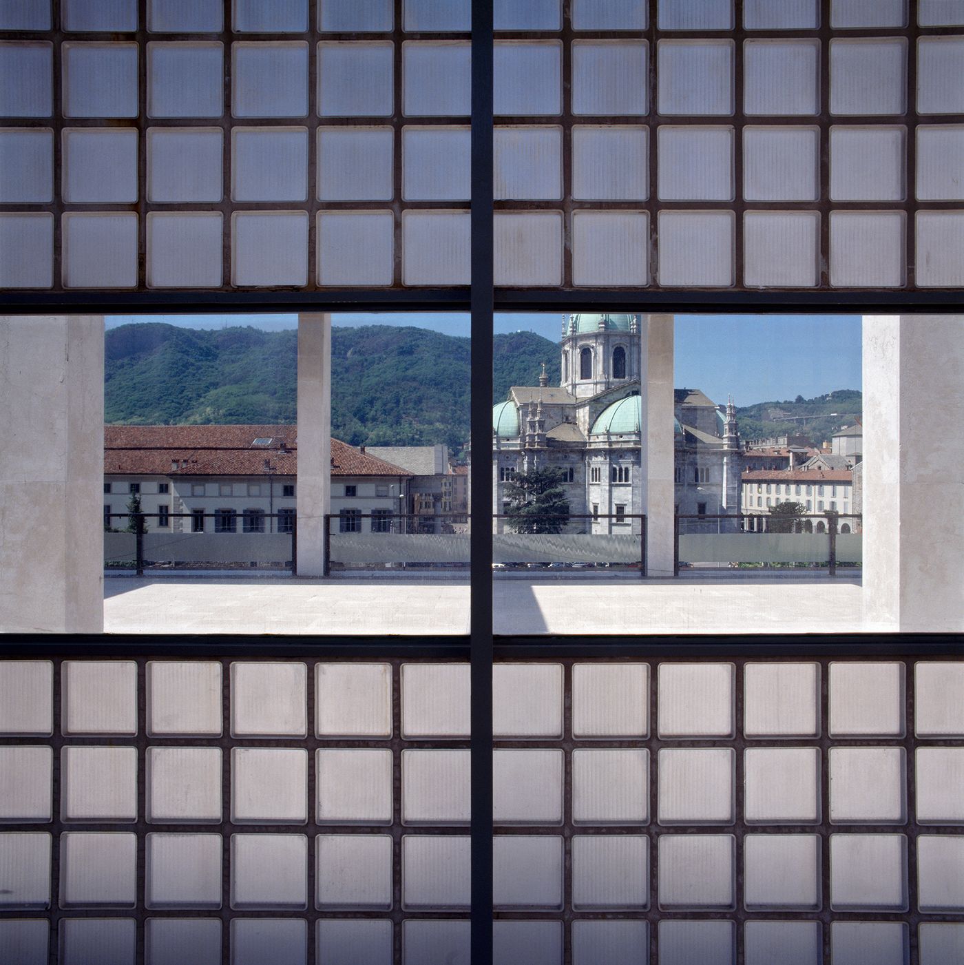 Viewed through a grid of glass blocks, a picturesque Italian cityscape unfolds. The foreground is defined by the textured grid of the window, while the background presents a charming scene of terracotta-tiled roofs and a grand domed church, set against rolling green hills under a clear blue sky. This framing emphasizes the interplay between interior architecture and the external environment, offering a glimpse of historic beauty through a contemporary lens.