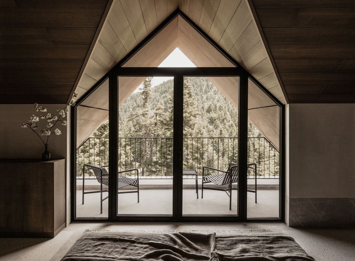 A captivating wide shot of a bedroom revealing a dramatic A-frame structure with dark timber framing and light wood paneling. The expansive window wall, leading to a balcony with modern black chairs, perfectly frames a breathtaking view of a dense, sun-drenched pine forest, emphasizing the seamless indoor-outdoor connection and a sense of peaceful retreat. A captivating wide shot of a bedroom revealing a dramatic A-frame structure with dark timber framing and light wood paneling. The expansive window wall, leading to a balcony with modern black chairs, perfectly frames a breathtaking view of a dense, sun-drenched pine forest, emphasizing the seamless indoor-outdoor connection and a sense of peaceful retreat.