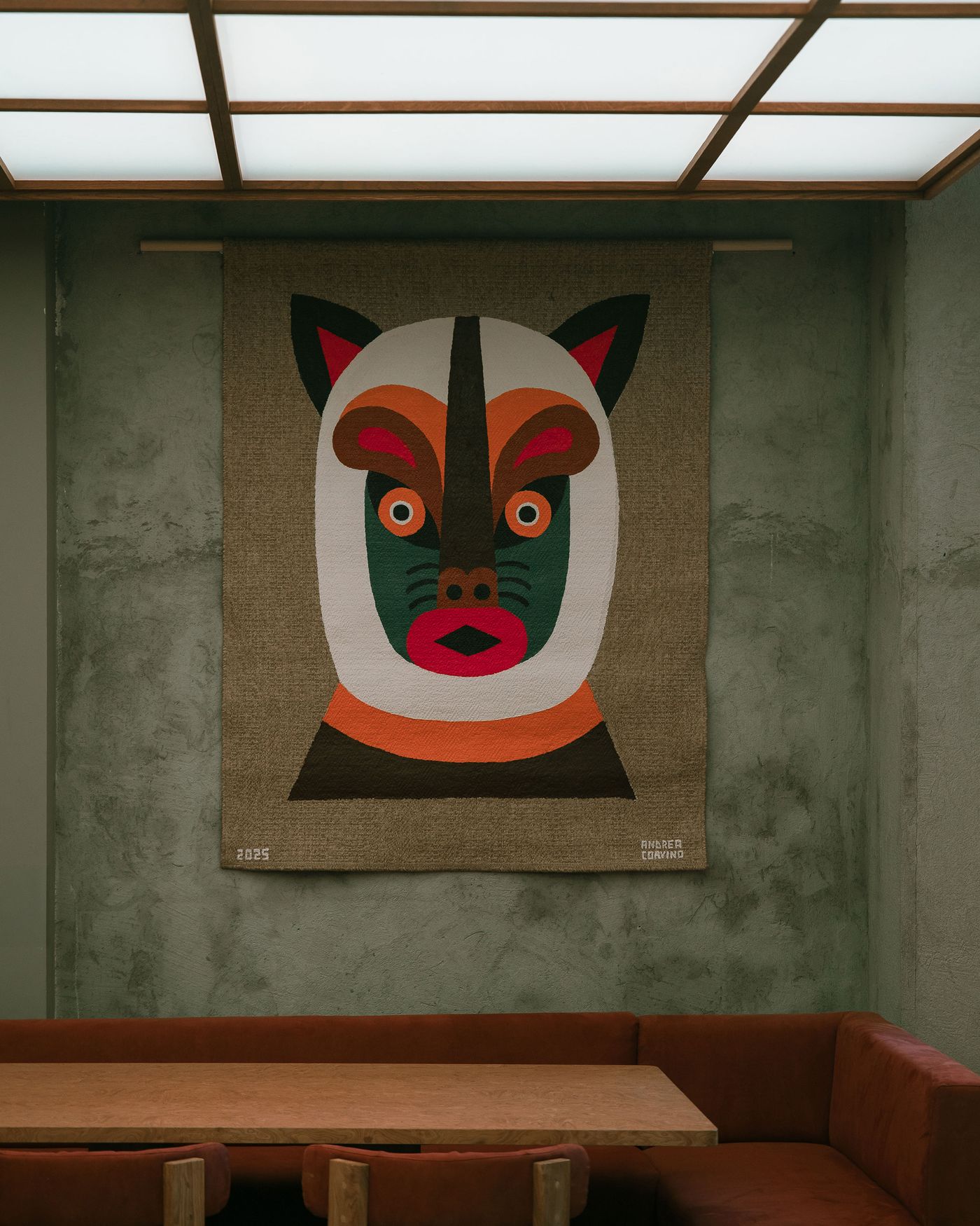 A colorful tapestry featuring a stylized animal face with bold geometric shapes, hanging on a textured wall above a wooden table and red seating. A colorful tapestry featuring a stylized animal face with bold geometric shapes, hanging on a textured wall above a wooden table and red seating.