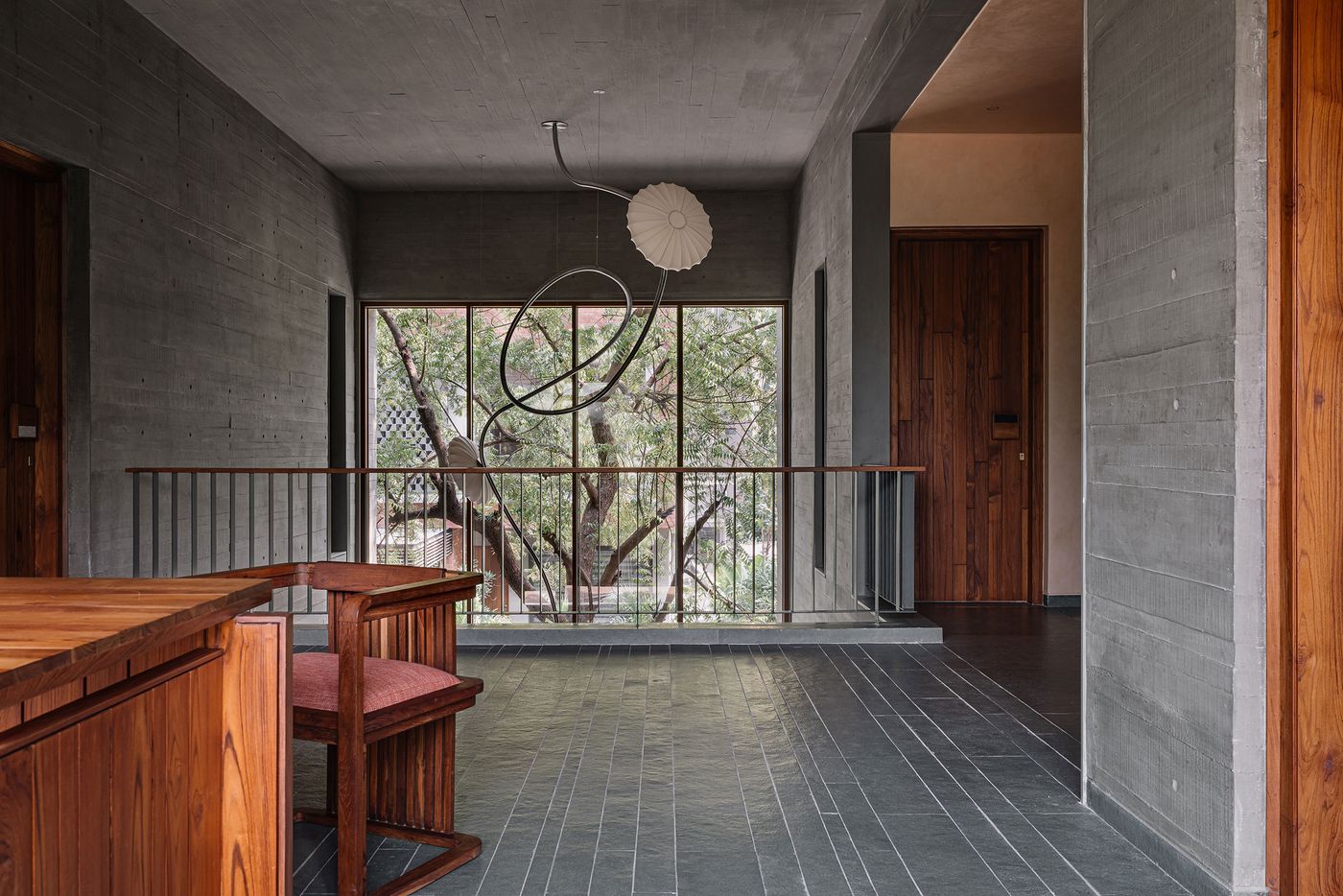 A broad interior view of a multi-level space with dark, textured concrete walls and a patterned dark grey tiled floor. A sculptural pendant light with organic, curved arms and a pleated shade hangs centrally. Expansive windows reveal a lush outdoor tree, and a minimalist metal railing delineates a lower level. Warm wooden furniture and doors add contrast and warmth.