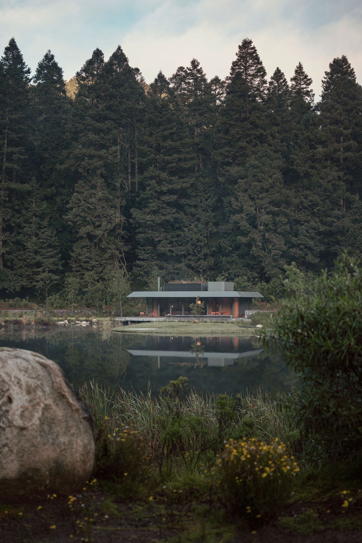 Modern house nestled by a serene lake, surrounded by lush evergreen forests, reflecting on the water's surface.