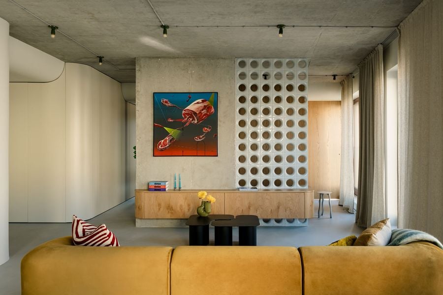 A bold artwork depicting surreal meat cuts floats against a raw concrete wall, flanked by a geometric plastic room divider. Below, a custom floating console in figured wood brings organic tactility, juxtaposed with minimalist black tables and playful styling details.
