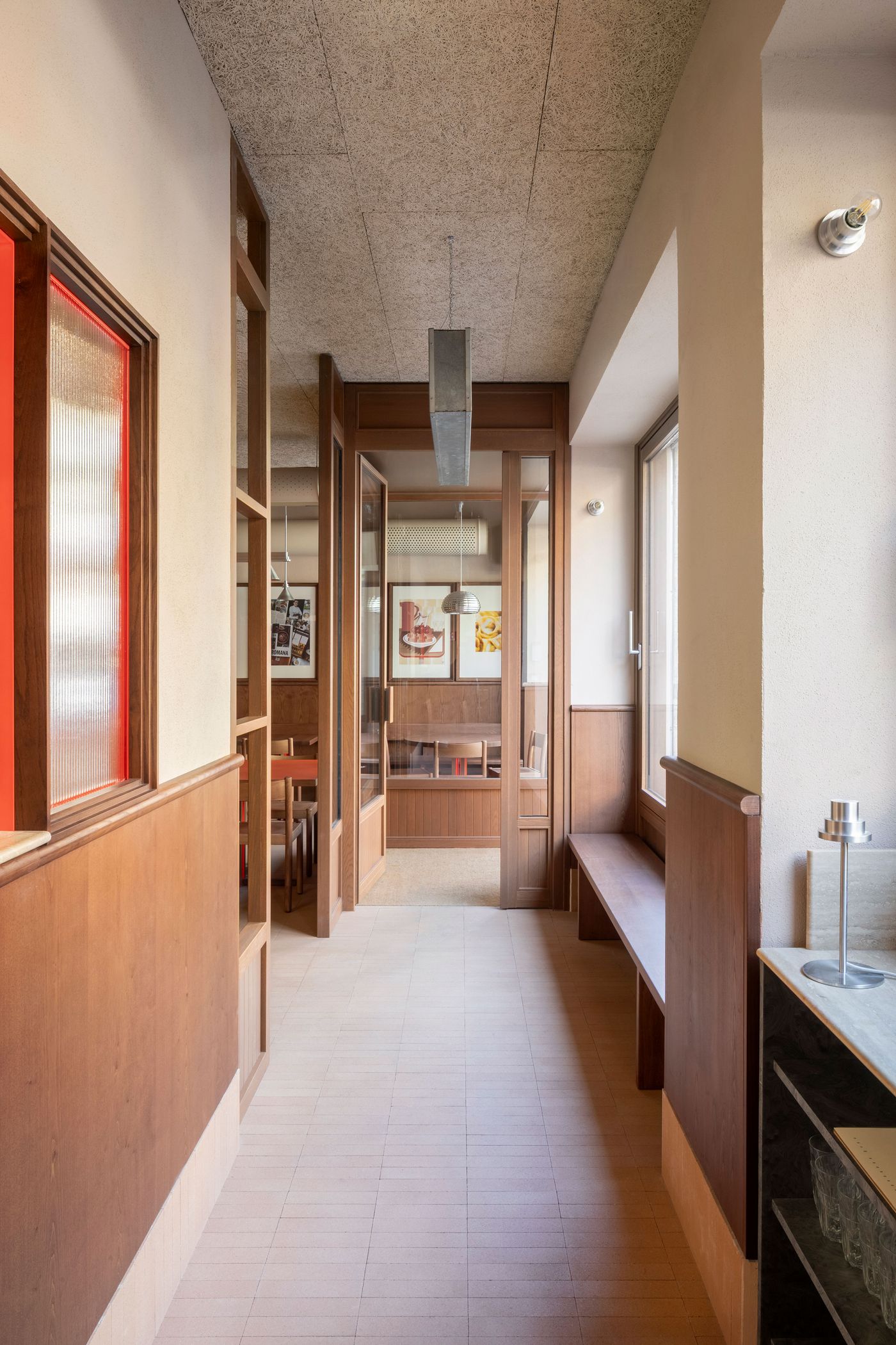 A warmly lit corridor at Santo Palato, designed by Naessi Studio, showcasing a refined neo-vernacular aesthetic. Wooden wainscoting and trim define the space, leading to a series of framed glass doors. A long wooden bench lines the right wall, while a striking red-framed window with fluted glass adds a pop of color and texture on the left. A warmly lit corridor at Santo Palato, designed by Naessi Studio, showcasing a refined neo-vernacular aesthetic. Wooden wainscoting and trim define the space, leading to a series of framed glass doors. A long wooden bench lines the right wall, while a striking red-framed window with fluted glass adds a pop of color and texture on the left.