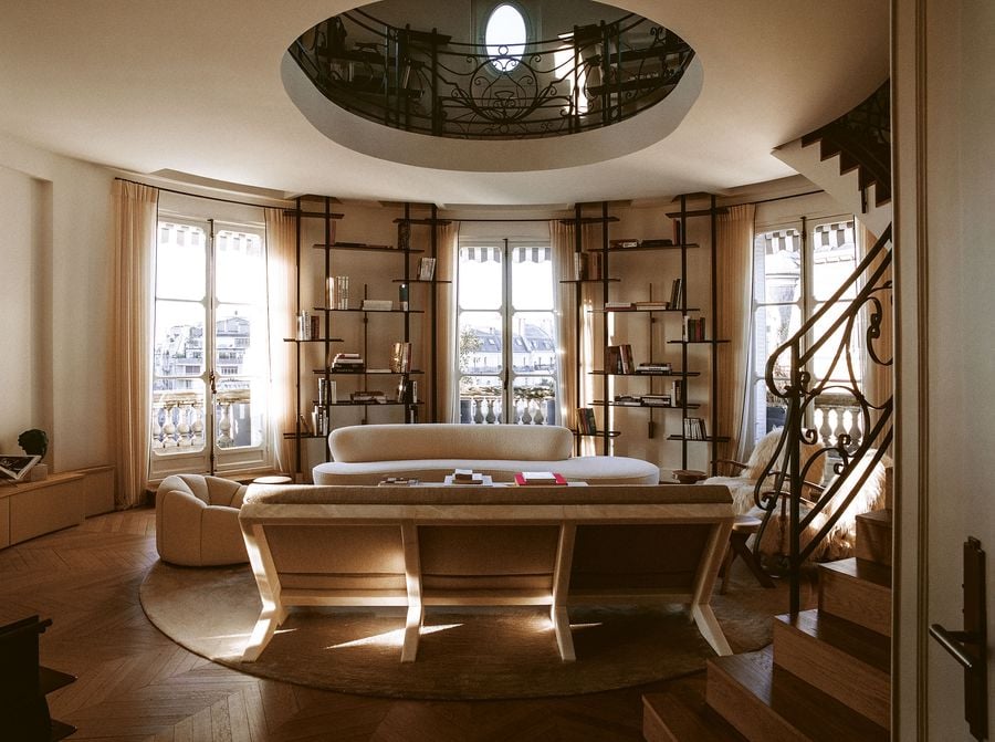 An opulent living area characterized by a grand elliptical opening in the ceiling, revealing a an intricate wrought-iron railing above. Curved modern sofas and a long, low console table define the seating arrangement, flanked by expansive windows offering city views, establishing a harmonious blend of traditional architecture and contemporary design.