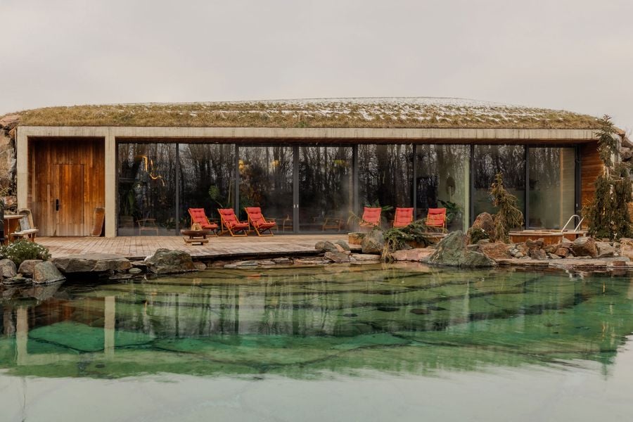 Modern eco-friendly house with a grassy roof, surrounded by a tranquil pond and red lounge chairs on a wooden deck.