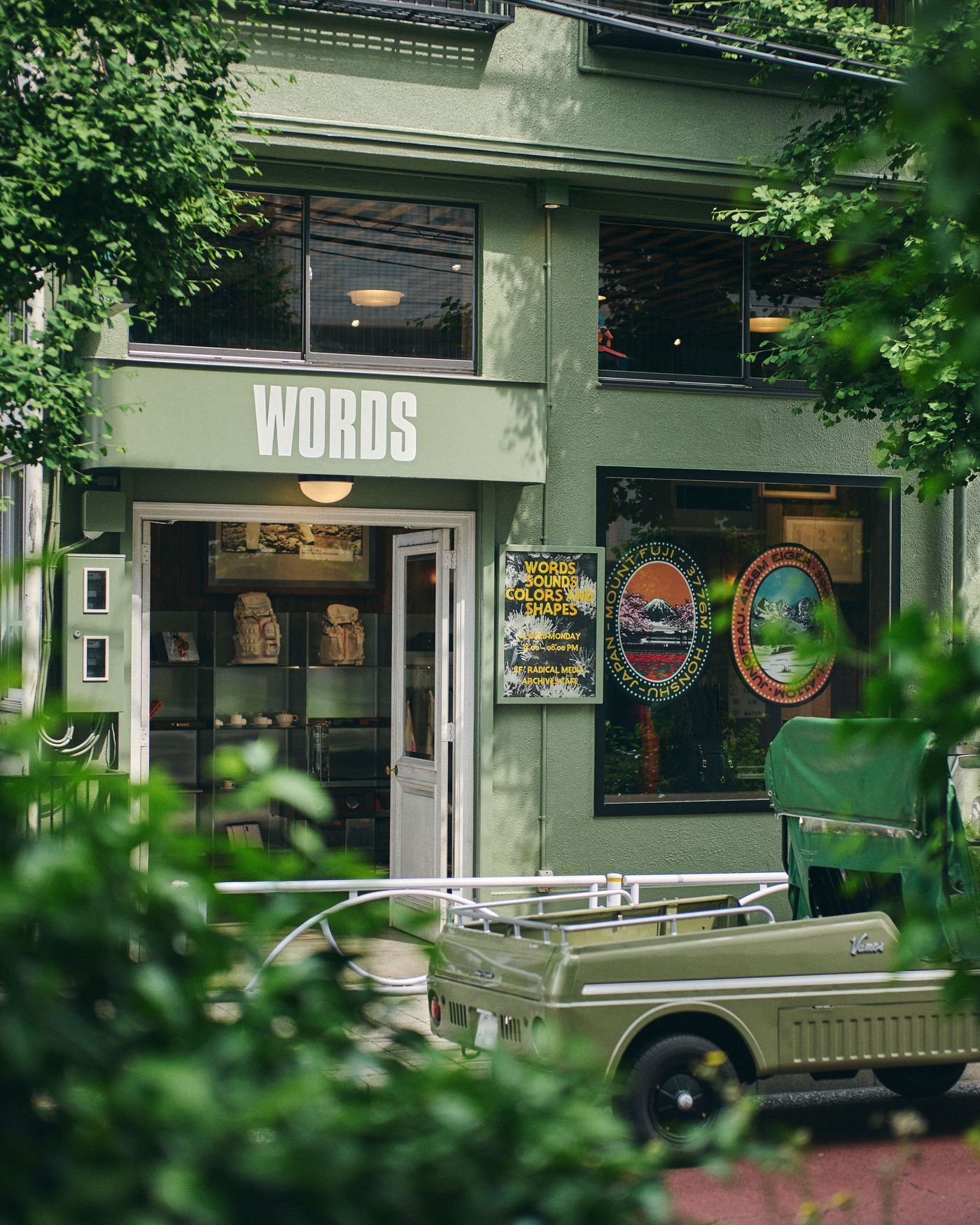A green storefront labeled "WORDS" displays art and posters, with a small vintage vehicle parked outside and lush foliage framing the scene.