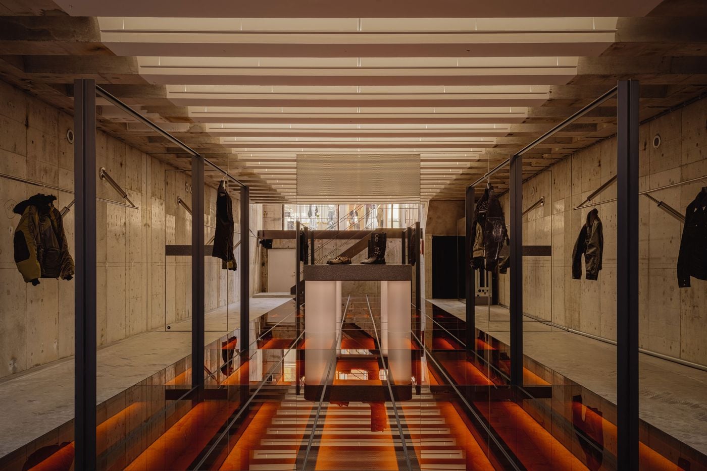 The basement is organised around a dramatic illuminated glass floor, glowing in deep red-orange beneath dark structural frames and display platforms. Above, closely spaced white ceiling elements establish a strong rhythm, while exposed concrete walls and sparse garment displays keep the composition austere, industrial and highly graphic.