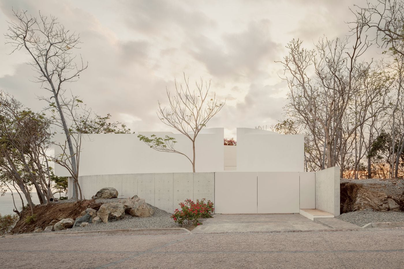 From the road, Casa Malonic withholds itself behind a curved concrete wall dotted with formwork ties, flush white gates and blank white volumes. Only a sliver of pink blossom in a narrow gap hints at life within, while boulders, grey gravel and leafless trees soften the threshold beneath a pale, clouded sky. From the road, Casa Malonic withholds itself behind a curved concrete wall dotted with formwork ties, flush white gates and blank white volumes. Only a sliver of pink blossom in a narrow gap hints at life within, while boulders, grey gravel and leafless trees soften the threshold beneath a pale, clouded sky.