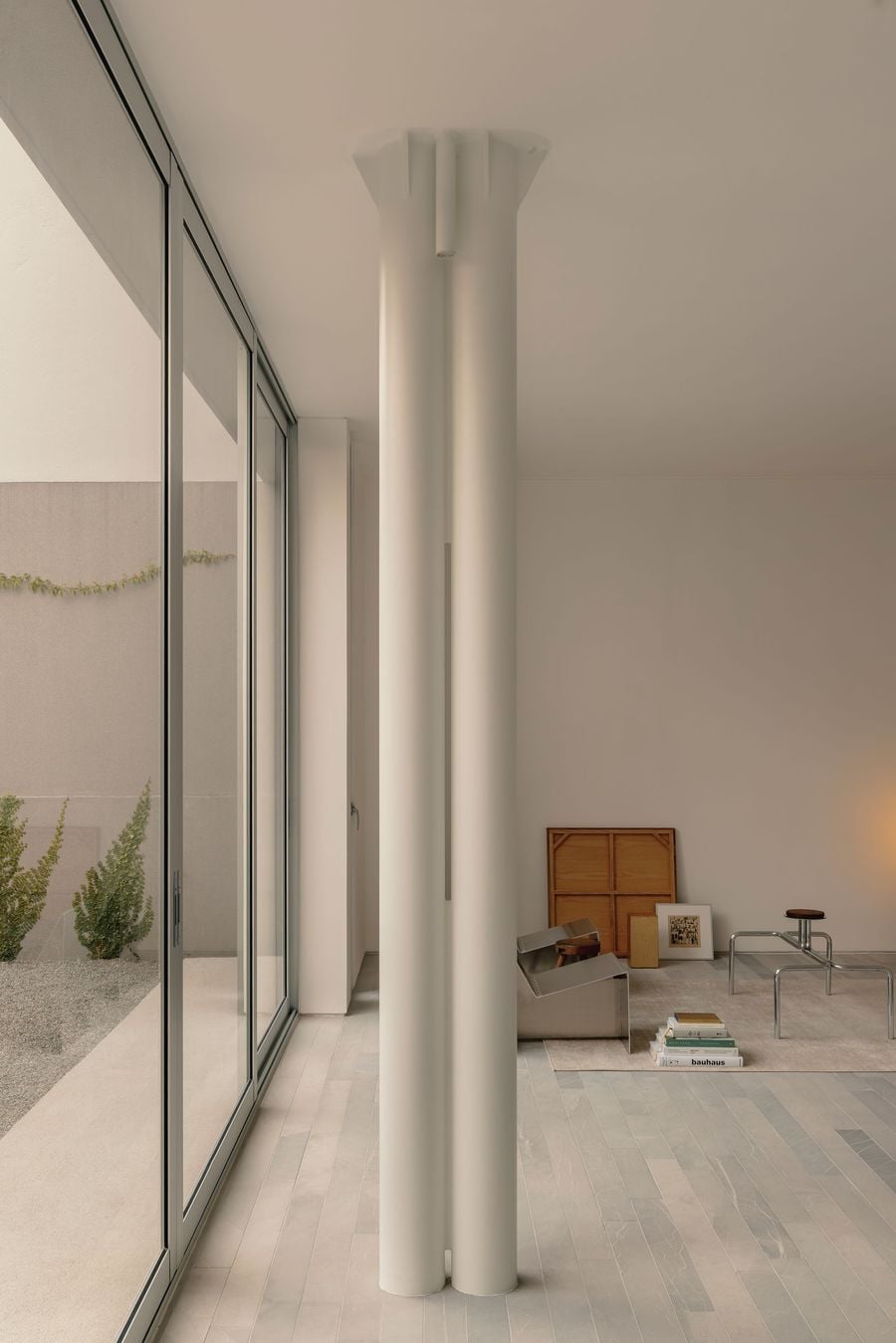 A cluster of slender white columns becomes an unexpectedly sculptural presence within Casa Tlaloc’s minimalist living room. Floor-to-ceiling glazing runs alongside the space, drawing in the courtyard greenery, while pale stone flooring, metal furniture, books and warm timber accents lend character to the otherwise pared-back interior. A cluster of slender white columns becomes an unexpectedly sculptural presence within Casa Tlaloc’s minimalist living room. Floor-to-ceiling glazing runs alongside the space, drawing in the courtyard greenery, while pale stone flooring, metal furniture, books and warm timber accents lend character to the otherwise pared-back interior.