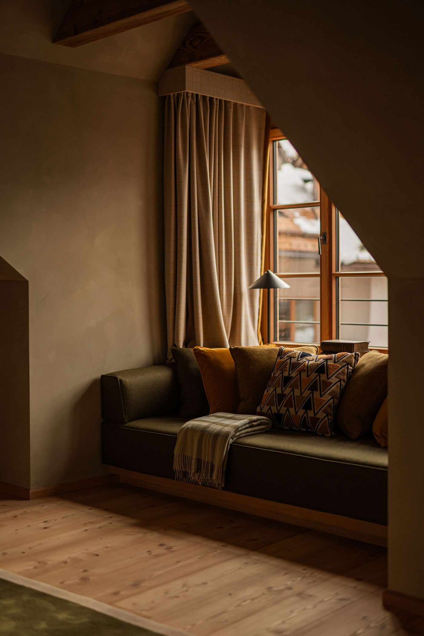 A built-in window seat fills a dormer niche at Castel Badia. It is upholstered in olive felted wool and piled with ochre velvet and geometric tufted cushions, with a fringed striped throw. Checked wool curtains, timber casement windows and pale wide-plank floors give the alcove a warm, cocooning quality. A built-in window seat fills a dormer niche at Castel Badia. It is upholstered in olive felted wool and piled with ochre velvet and geometric tufted cushions, with a fringed striped throw. Checked wool curtains, timber casement windows and pale wide-plank floors give the alcove a warm, cocooning quality.