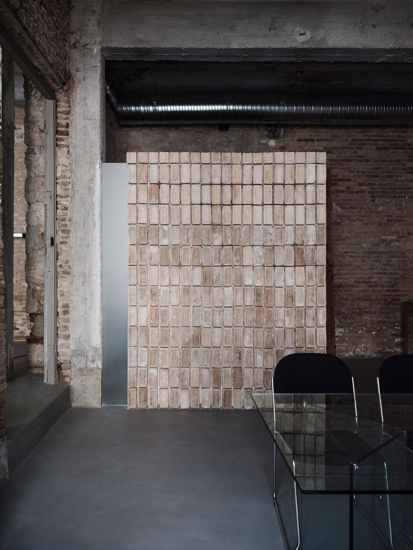 Reclaimed bricks form a freestanding partition behind a glass meeting table and black tubular-steel chairs. Their warm, uneven surfaces contrast with the smooth grey floor, exposed ductwork and raw masonry surrounding them, demonstrating how salvaged material has been reassembled into purposeful new elements without disguising its age. Reclaimed bricks form a freestanding partition behind a glass meeting table and black tubular-steel chairs. Their warm, uneven surfaces contrast with the smooth grey floor, exposed ductwork and raw masonry surrounding them, demonstrating how salvaged material has been reassembled into purposeful new elements without disguising its age.