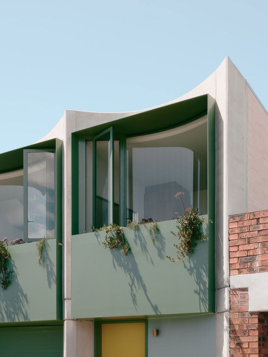 A close-up of Denj Lane’s sculptural upper façade reveals the sweeping concave concrete profile and deep sage-green window bay. Operable glazing and cascading succulents soften the robust materials, while strong sunlight casts elongated botanical shadows across the façade, reinforcing the project’s interplay of architecture, landscape and colour. A close-up of Denj Lane’s sculptural upper façade reveals the sweeping concave concrete profile and deep sage-green window bay. Operable glazing and cascading succulents soften the robust materials, while strong sunlight casts elongated botanical shadows across the façade, reinforcing the project’s interplay of architecture, landscape and colour.