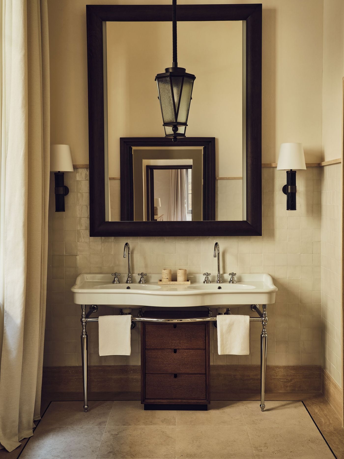 A double console basin on polished nickel legs stands against glazed, hand-finished tiles in a bathroom at La Réserve Firenze. A lantern pendant hangs before a large dark-framed mirror whose reflection opens onto an enfilade of doorways, while a stone floor and linen curtain keep the palette soft and luminous. A double console basin on polished nickel legs stands against glazed, hand-finished tiles in a bathroom at La Réserve Firenze. A lantern pendant hangs before a large dark-framed mirror whose reflection opens onto an enfilade of doorways, while a stone floor and linen curtain keep the palette soft and luminous.