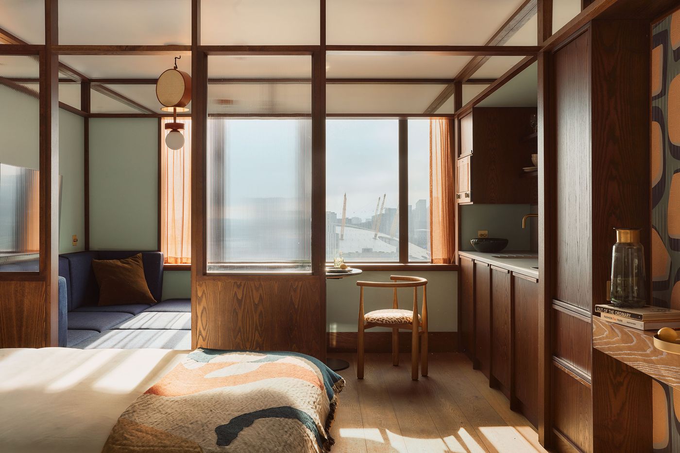 A guest room pairs sage-green walls and oak flooring with a rigorous grid of dark walnut framing that extends across walls and ceiling. Reeded-glass partitions delineate the sleeping, living and kitchen areas while preserving light and openness, with broad windows framing views towards the Thames and Greenwich Peninsula. A guest room pairs sage-green walls and oak flooring with a rigorous grid of dark walnut framing that extends across walls and ceiling. Reeded-glass partitions delineate the sleeping, living and kitchen areas while preserving light and openness, with broad windows framing views towards the Thames and Greenwich Peninsula.