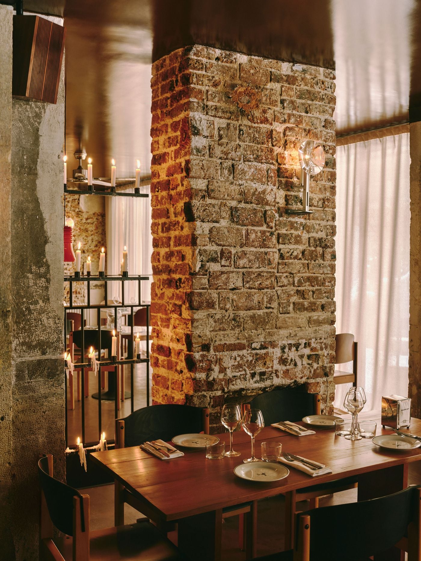A timber table set for dinner nestles against the base of a rugged brick pillar at SaltoBar in Paris. A glass sconce catches the light on the weathered masonry. To the left, tiers of dripping candles on a black metal screen glow against raw concrete. Sheer curtains diffuse the daylight behind. A timber table set for dinner nestles against the base of a rugged brick pillar at SaltoBar in Paris. A glass sconce catches the light on the weathered masonry. To the left, tiers of dripping candles on a black metal screen glow against raw concrete. Sheer curtains diffuse the daylight behind.