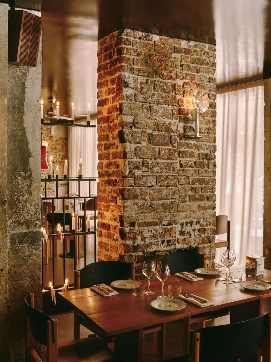 A timber table set for dinner nestles against the base of a rugged brick pillar at SaltoBar in Paris. A glass sconce catches the light on the weathered masonry. To the left, tiers of dripping candles on a black metal screen glow against raw concrete. Sheer curtains diffuse the daylight behind. A timber table set for dinner nestles against the base of a rugged brick pillar at SaltoBar in Paris. A glass sconce catches the light on the weathered masonry. To the left, tiers of dripping candles on a black metal screen glow against raw concrete. Sheer curtains diffuse the daylight behind.