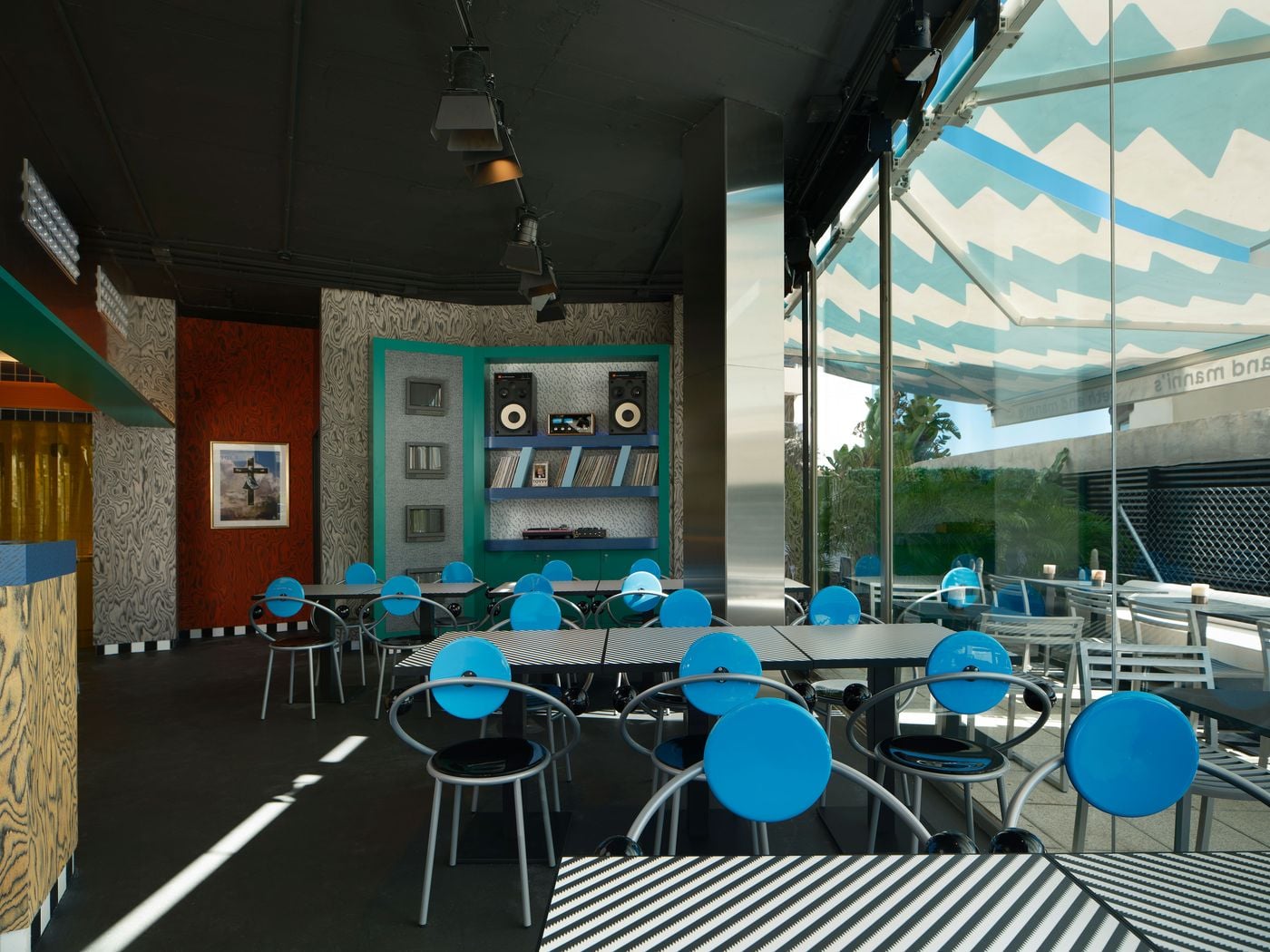 In the dining room of Seth & Manni's, rows of Michele De Lucchi's First chairs gather around striped tables, their glossy blue disc backs and black spheres catching the light. A teal unit of speakers, records and monitors stands against grey wood-grain walls. A full-height glass wall opens onto the terrace beneath a zigzag awning. In the dining room of Seth & Manni's, rows of Michele De Lucchi's First chairs gather around striped tables, their glossy blue disc backs and black spheres catching the light. A teal unit of speakers, records and monitors stands against grey wood-grain walls. A full-height glass wall opens onto the terrace beneath a zigzag awning.