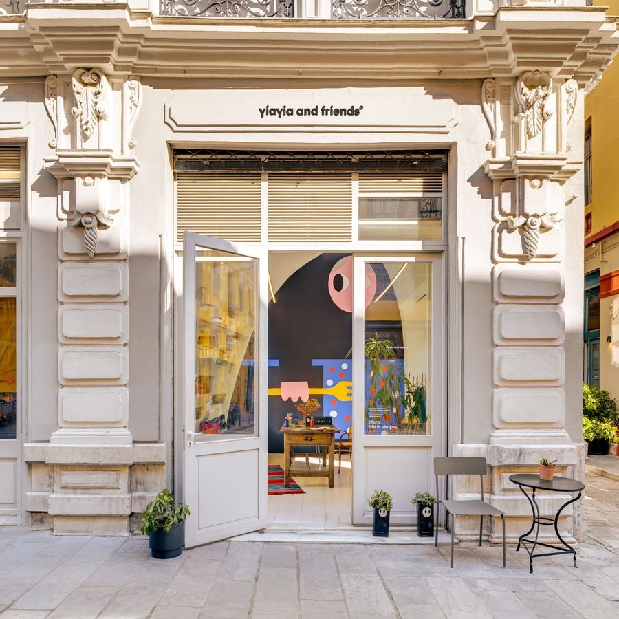 Yiayia and Friends storefront in a restored neoclassical building with carved stone brackets and rusticated pilasters, open glass doors revealing a colorful graphic mural, a rustic wooden table and kilim rug, with potted plants on the marble pavement.