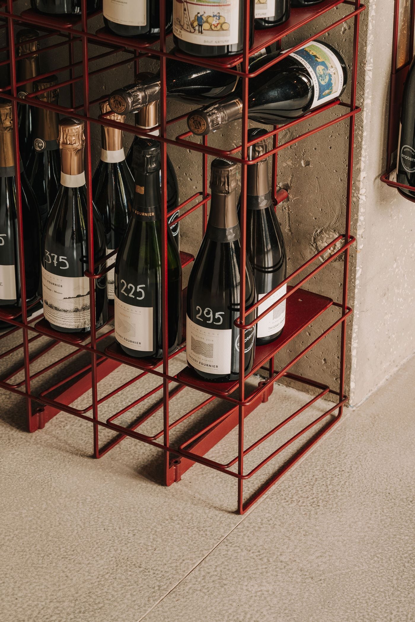 A close-up of WIN's custom wine shelving reveals an intricate framework of slender burgundy metal rods, with individual compartments accommodating champagne bottles both upright and horizontally. The precise geometry of the structure contrasts with the rough concrete wall behind it and the pale, subtly speckled flooring below. A close-up of WIN's custom wine shelving reveals an intricate framework of slender burgundy metal rods, with individual compartments accommodating champagne bottles both upright and horizontally. The precise geometry of the structure contrasts with the rough concrete wall behind it and the pale, subtly speckled flooring below.