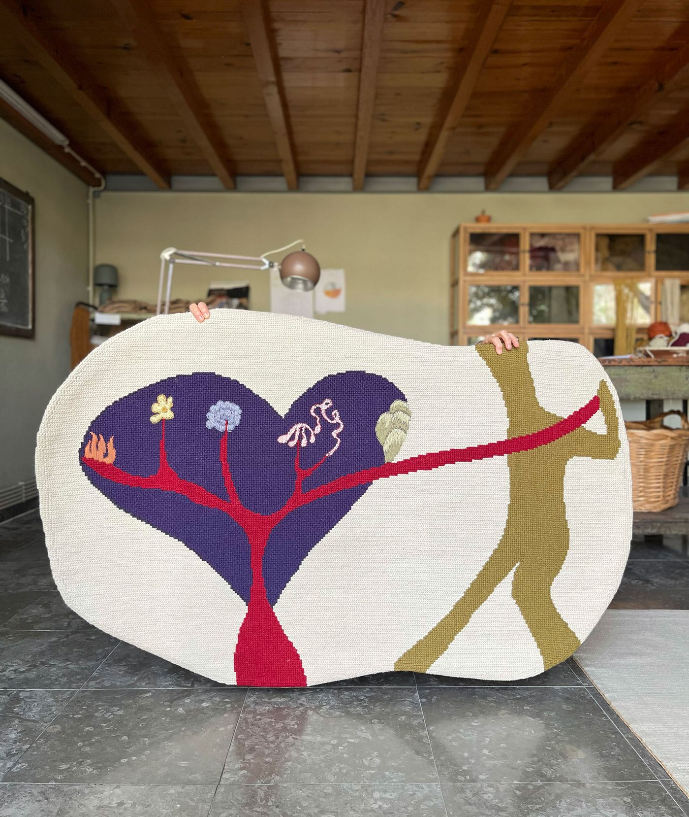 Two hands hold up a large, irregularly shaped rug by Defio Rugs, depicting a whimsical, abstract scene. On a cream background, a large purple heart-shaped tree with red roots and small blue, yellow, and green flowers intertwines with a stylized olive-green human figure. A vibrant red ribbon connects the two, symbolizing connection and vitality. Two hands hold up a large, irregularly shaped rug by Defio Rugs, depicting a whimsical, abstract scene. On a cream background, a large purple heart-shaped tree with red roots and small blue, yellow, and green flowers intertwines with a stylized olive-green human figure. A vibrant red ribbon connects the two, symbolizing connection and vitality.