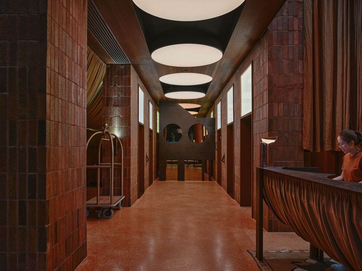 A stylish hotel lobby featuring warm-toned brick walls, round ceiling lights, and a reception desk with a luggage cart nearby. A stylish hotel lobby featuring warm-toned brick walls, round ceiling lights, and a reception desk with a luggage cart nearby.