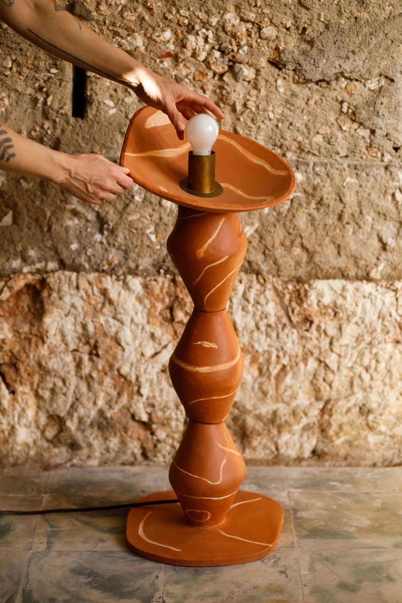 Hands adjust a light bulb on a unique terracotta-colored floor lamp by Pareidolia Design, set against a rustic stone wall. The lamp features a stacked, organic, hourglass-like base with linear white markings, topped by a shallow, curved shade. The raw, handcrafted aesthetic of the lamp contrasts beautifully with the rugged texture of the wall.