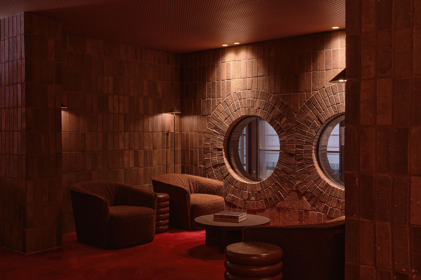 Cozy, dimly lit lounge with textured brown walls, circular windows, plush chairs, and a round table, exuding warmth and style.