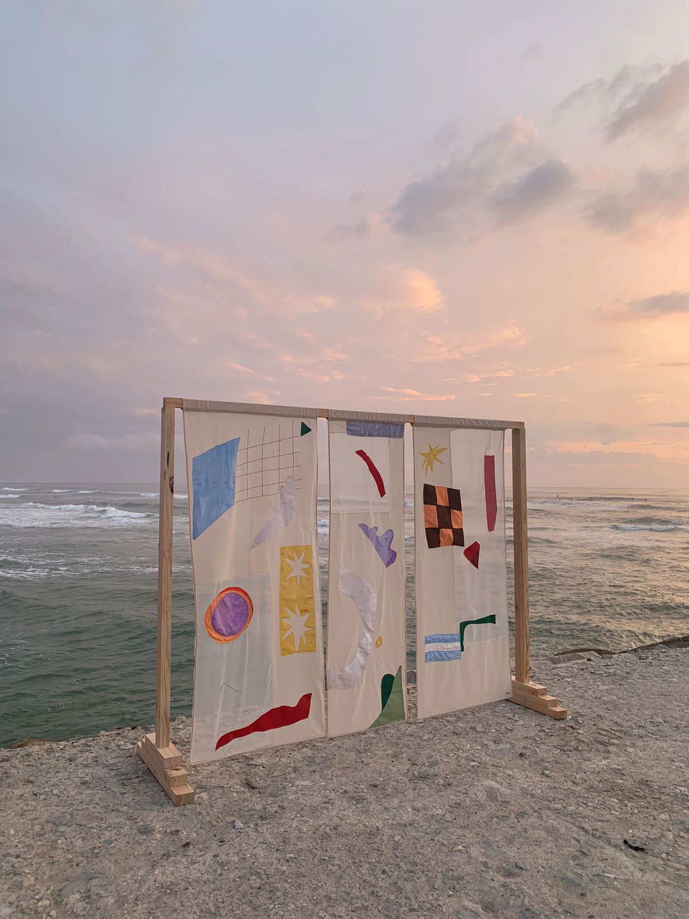A striking textile art installation by Maria Dezasseis, featuring three vertical panels of light-colored fabric adorned with colorful abstract shapes in blue, purple, red, and yellow. The panels are framed by a simple wooden structure, set against a serene backdrop of a cloudy sky and calm ocean waves, blending artistic expression with natural beauty.