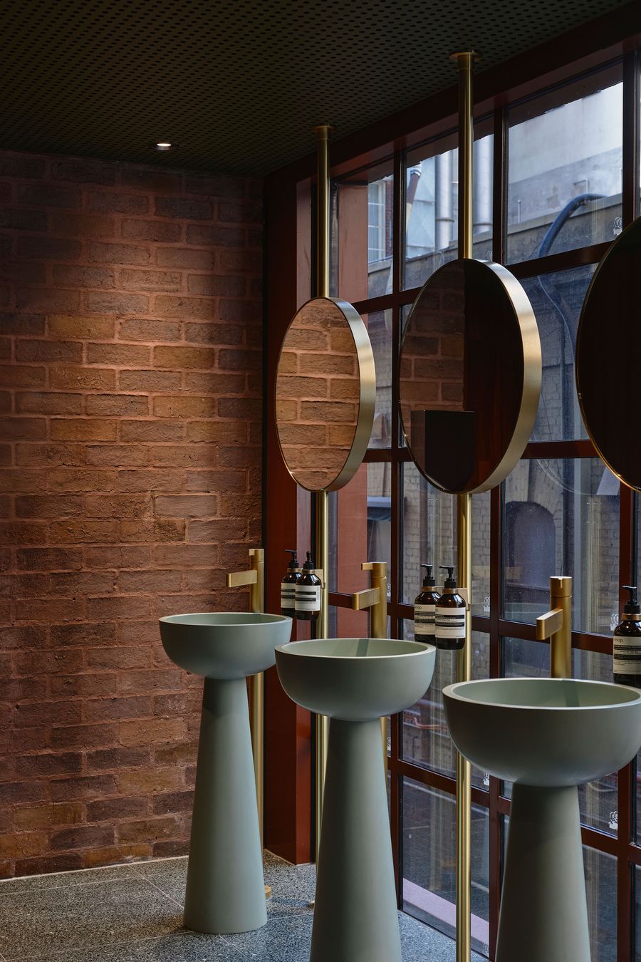 Modern restroom with three pastel green basins, gold mirrors, and brick walls, featuring stylish soap dispensers beside large windows. Modern restroom with three pastel green basins, gold mirrors, and brick walls, featuring stylish soap dispensers beside large windows.