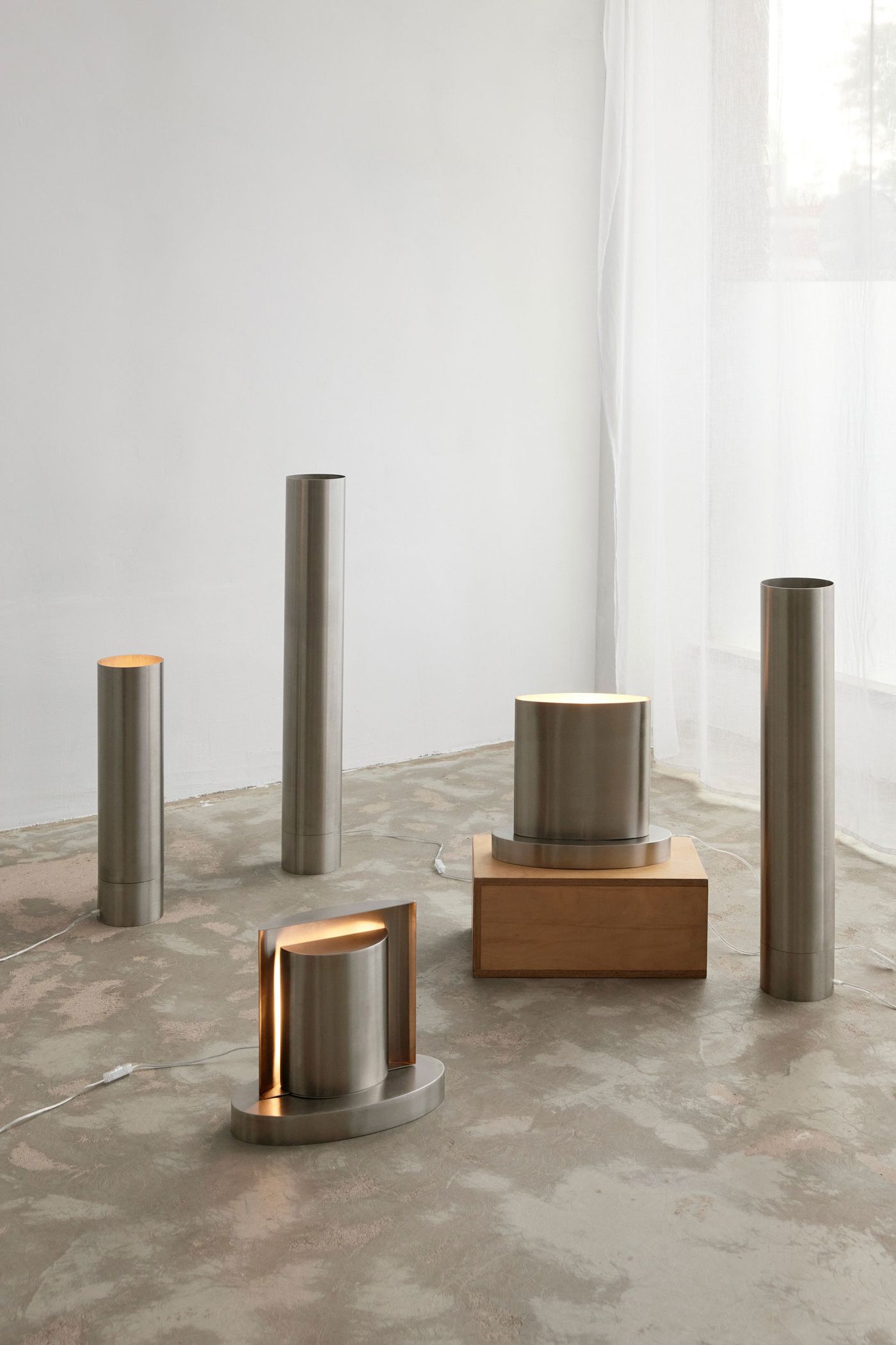 A minimalist collection of five cylindrical and rectangular lamps by Thayra Correia, crafted from brushed metal and wood, is artfully arranged on a polished concrete floor. The subtle illumination from within these sculptural forms creates a serene atmosphere, highlighting their clean lines and understated industrial-meets-Japandi aesthetic. A minimalist collection of five cylindrical and rectangular lamps by Thayra Correia, crafted from brushed metal and wood, is artfully arranged on a polished concrete floor. The subtle illumination from within these sculptural forms creates a serene atmosphere, highlighting their clean lines and understated industrial-meets-Japandi aesthetic.