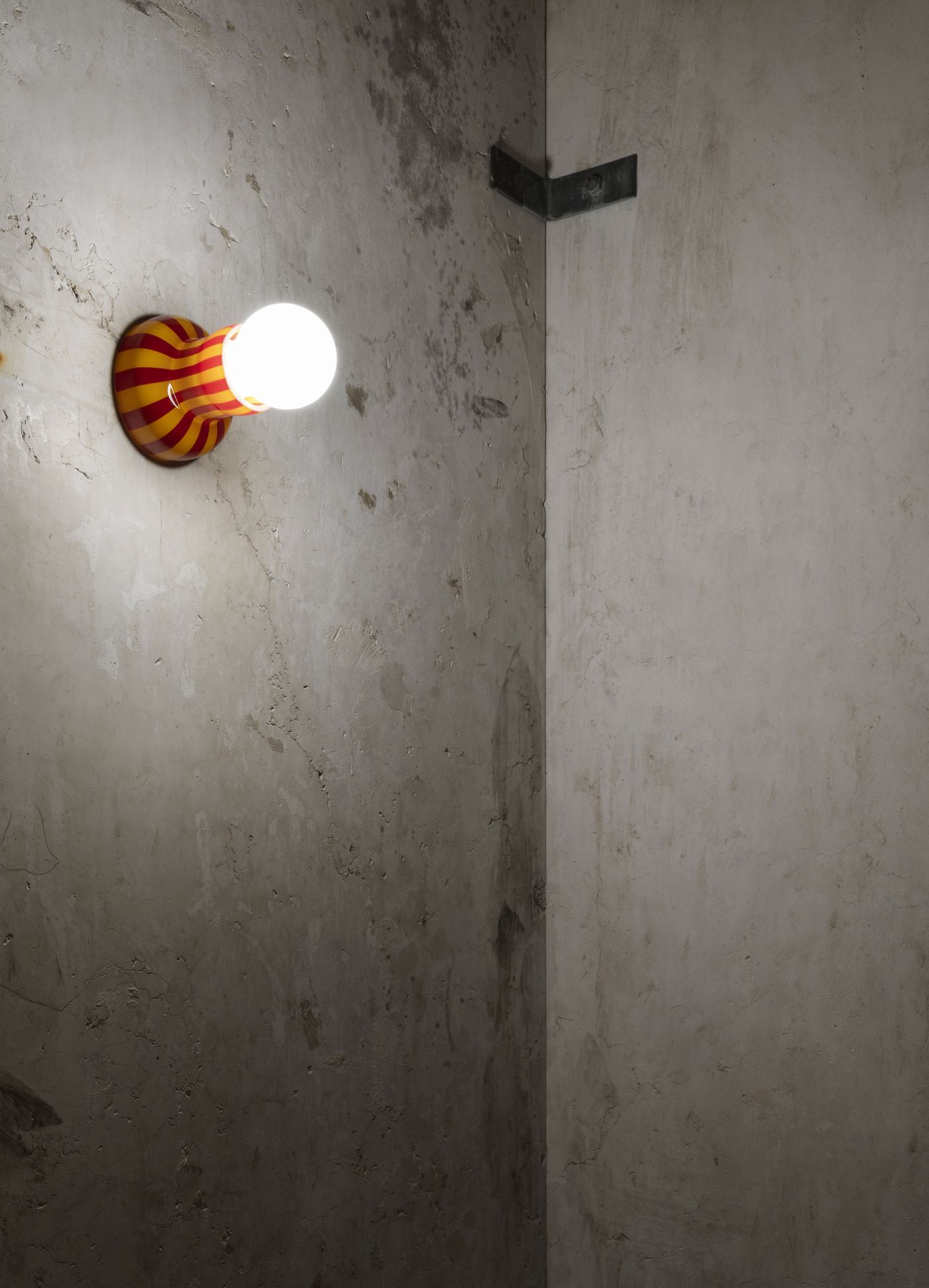 A playful wall light with red and yellow stripes brightens a textured, raw concrete corner.