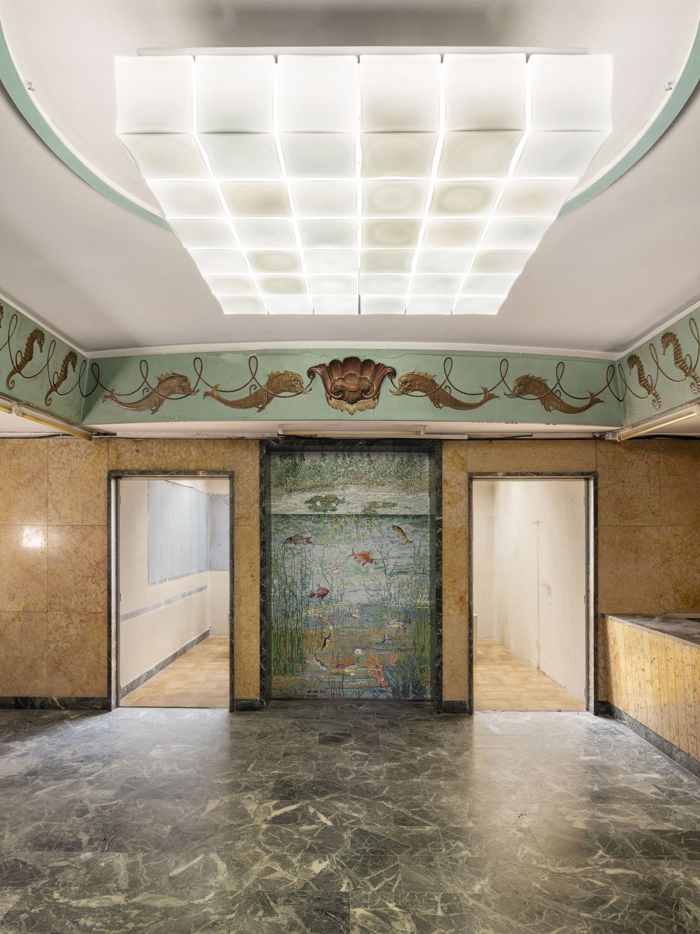 A grand Art Deco interior with green marble flooring, fish and shell motifs along a pale green frieze, and a vivid mosaic depicting aquatic life between two doorways beneath a modern square-paneled ceiling light.
