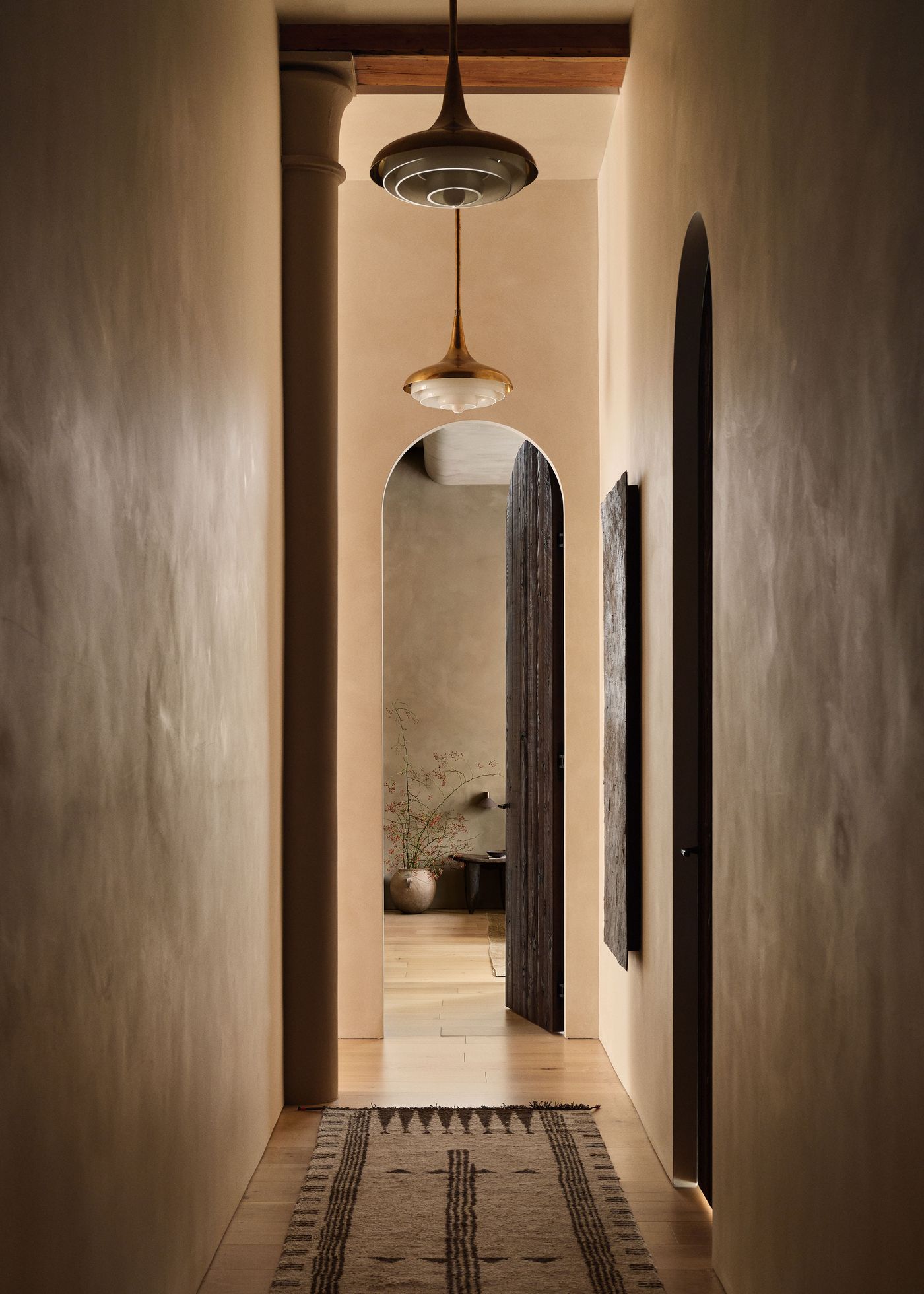 A softly lit hallway with arched doorways, warm beige walls, wooden flooring, and stylish light fixtures, leading to a cozy room.