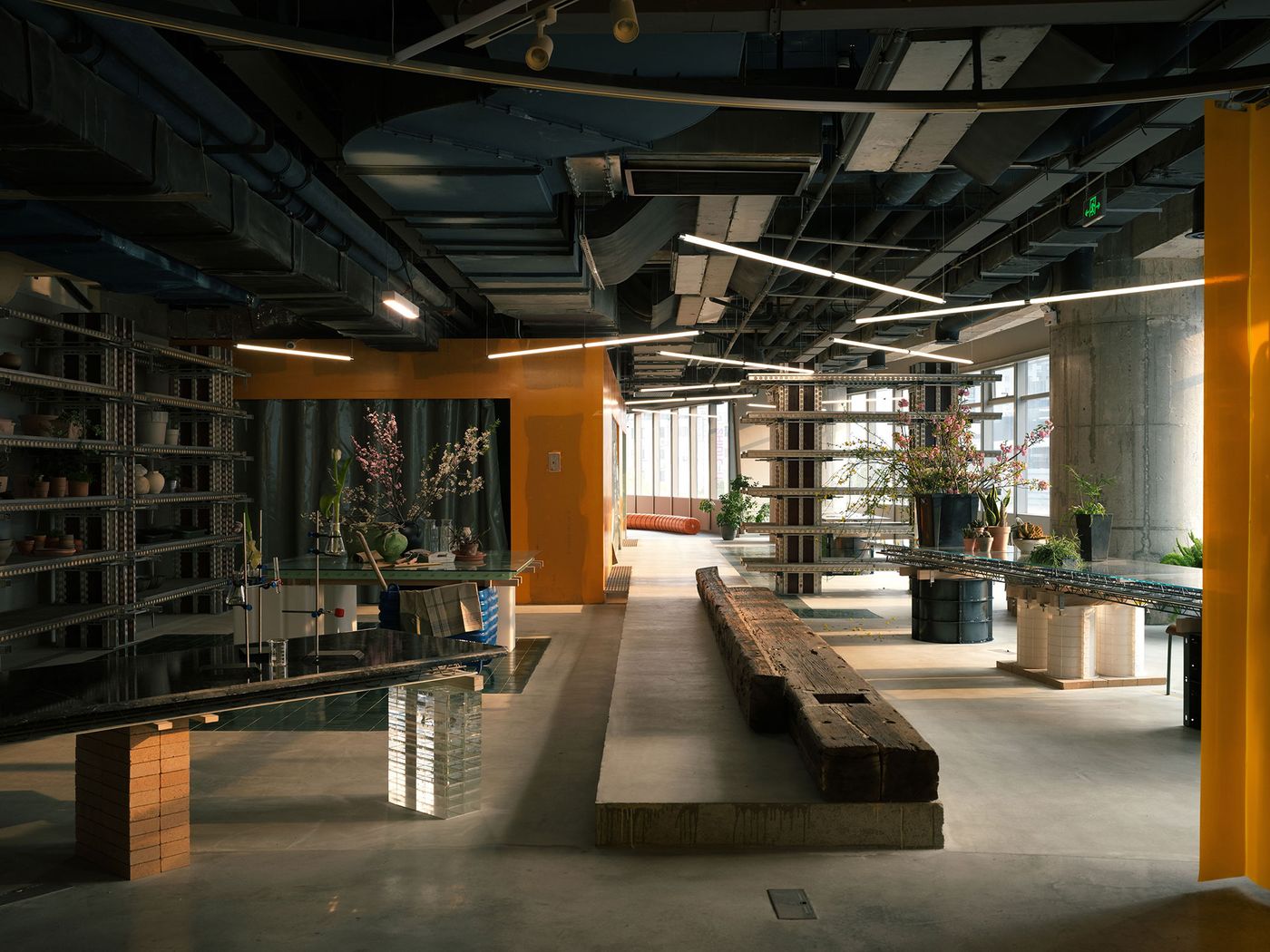 This expansive view of 889 GLO Art Space highlights its industrial architecture with exposed ceilings and concrete elements. Long, minimalist tables, some with a dark, reflective surface, are paired with a rustic wooden bench. Vibrant yellow partitions add warmth, defining zones within the vast, multi-functional space, reflecting a blend of brutalist and modern industrial aesthetics. This expansive view of 889 GLO Art Space highlights its industrial architecture with exposed ceilings and concrete elements. Long, minimalist tables, some with a dark, reflective surface, are paired with a rustic wooden bench. Vibrant yellow partitions add warmth, defining zones within the vast, multi-functional space, reflecting a blend of brutalist and modern industrial aesthetics.