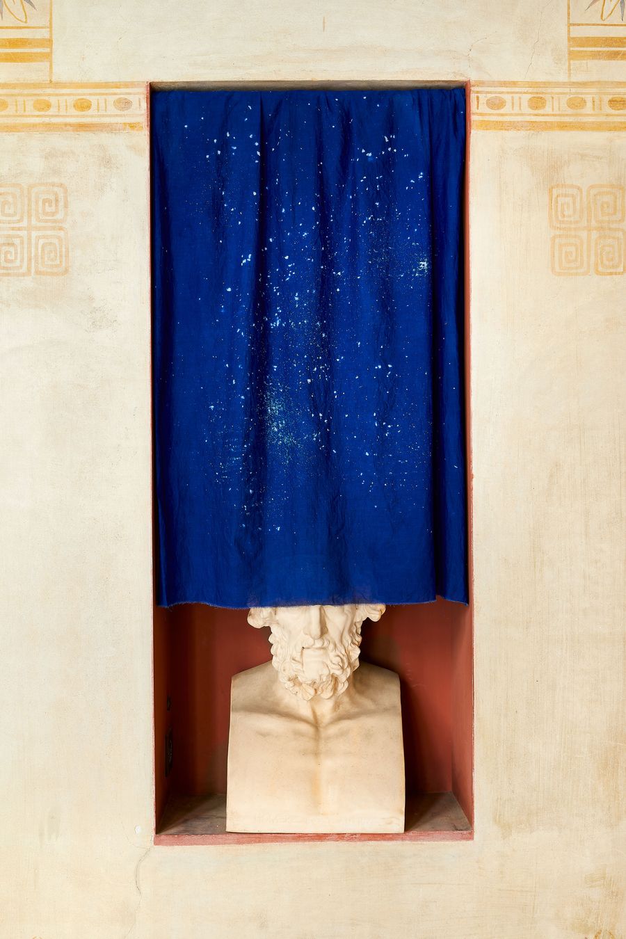 A tight, intimate shot of a marble bust of a bearded man, set within a red-hued niche. A curtain of deep, cobalt blue hangs from the top of the alcove, splattered with white spots. The simple composition highlights the blend of classical form and modern, minimalist interruption. A tight, intimate shot of a marble bust of a bearded man, set within a red-hued niche. A curtain of deep, cobalt blue hangs from the top of the alcove, splattered with white spots. The simple composition highlights the blend of classical form and modern, minimalist interruption.