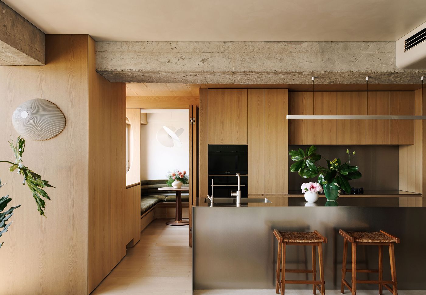 An open-plan kitchen and dining area in a Shanghai apartment designed by YSP Design, showcasing a harmonious blend of industrial and Japandi styles. The kitchen features sleek wooden cabinetry and a brushed stainless steel island, while a cozy dining nook with olive green seating is visible through an open doorway. Exposed concrete beams add a raw, industrial edge. An open-plan kitchen and dining area in a Shanghai apartment designed by YSP Design, showcasing a harmonious blend of industrial and Japandi styles. The kitchen features sleek wooden cabinetry and a brushed stainless steel island, while a cozy dining nook with olive green seating is visible through an open doorway. Exposed concrete beams add a raw, industrial edge.