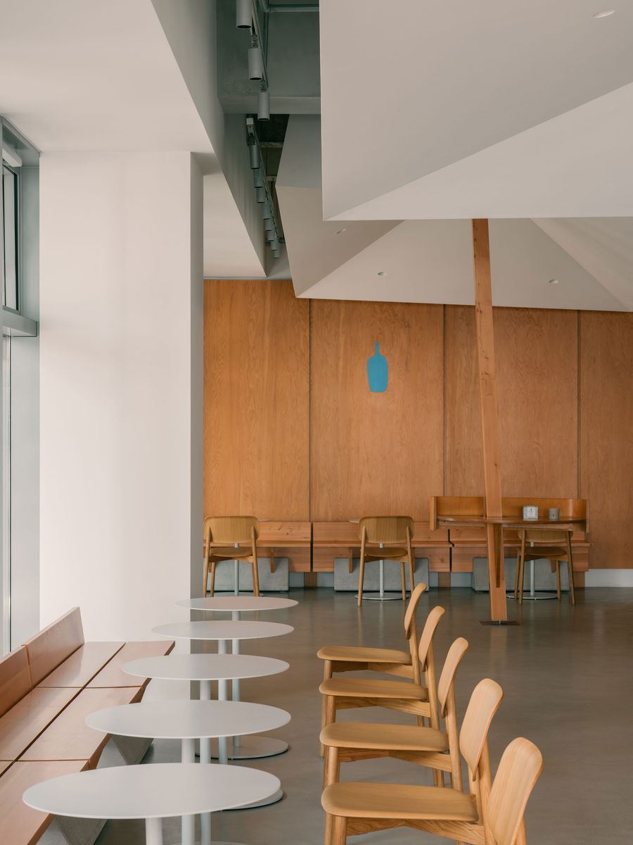 Linear arrangements of light wood chairs and oval café tables mirror the rhythm of the architectural ceiling planes, culminating in a Blue Bottle logo subtly painted on the oak-paneled wall.