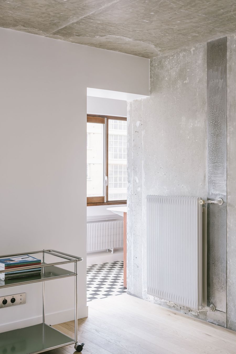 Modern interior featuring a concrete wall, a white radiator, a glass-topped cart, and a visible patterned floor through an open doorway. Modern interior featuring a concrete wall, a white radiator, a glass-topped cart, and a visible patterned floor through an open doorway.