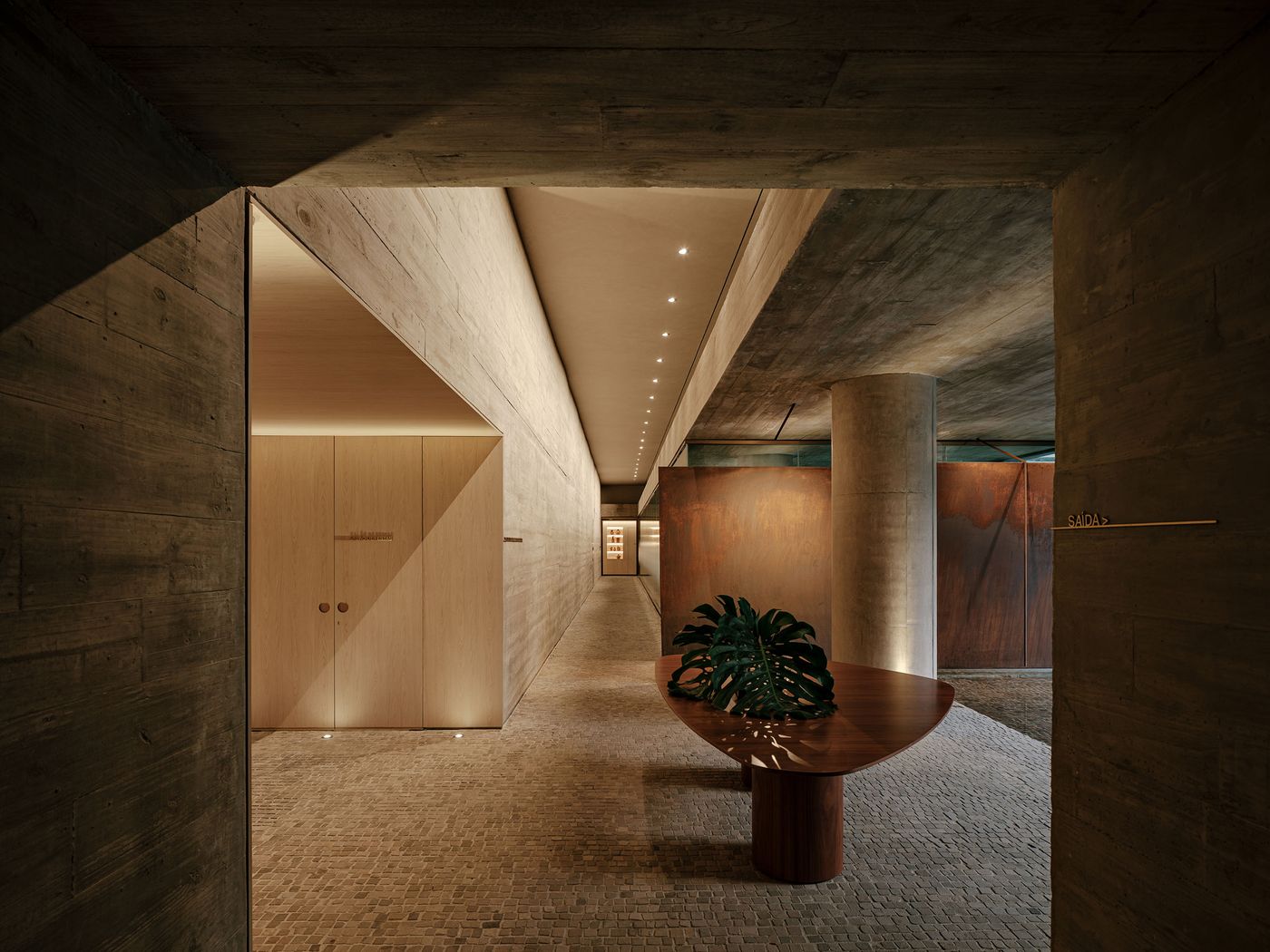 A sophisticated, low-lit hallway, characterized by a play of light and shadow on raw concrete and warm wooden surfaces. A sculptural half-moon console table with a large green plant adds organic form, while subtle linear lighting guides the eye through the meticulously crafted space. A sophisticated, low-lit hallway, characterized by a play of light and shadow on raw concrete and warm wooden surfaces. A sculptural half-moon console table with a large green plant adds organic form, while subtle linear lighting guides the eye through the meticulously crafted space.