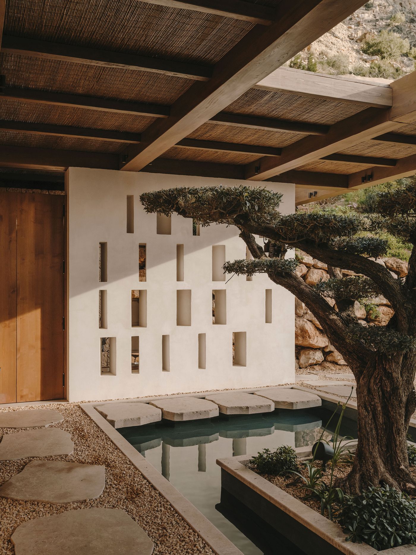 A serene courtyard featuring a minimalist design, with a textured wall, stepping stones over water, and a lush bonsai tree.