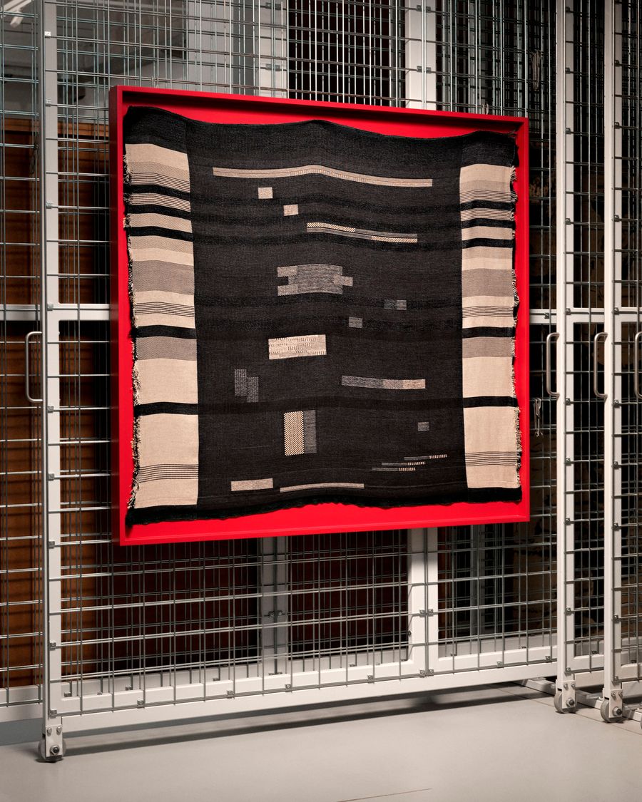 A striking woven textile, predominantly black with intricate bands of cream and grey geometric patterns, is centrally displayed within a vivid red frame. This artwork is suspended against a background of industrial wire grids and metal shelving. The stark contrast between the textured fabric, the bold frame, and the utilitarian grid structure creates a dynamic visual tension, hinting at an exhibition of craft or design within a modern, minimalist setting.