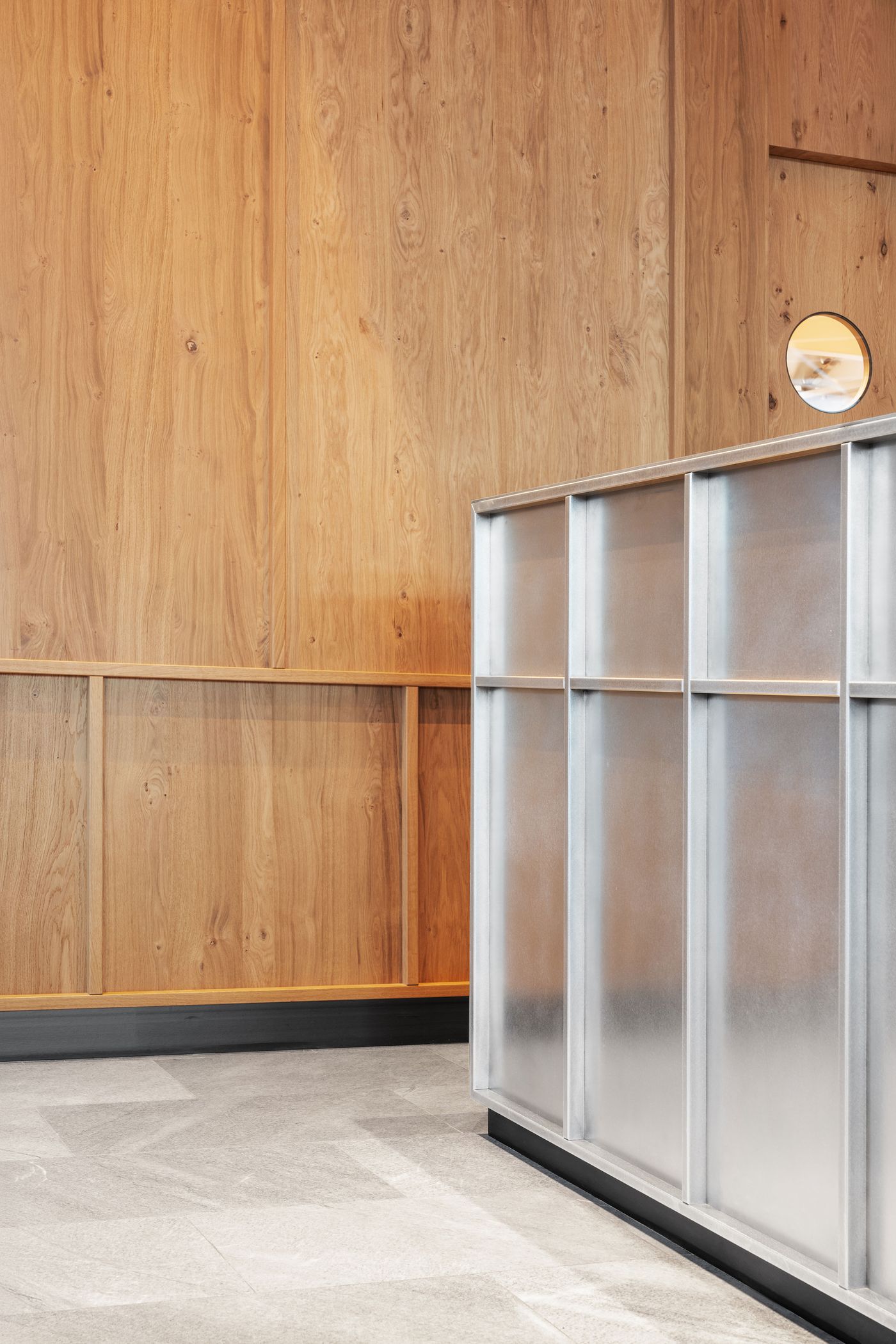 Floor-to-ceiling timber paneling showcases natural wood grain patterns alongside sleek steel display elements. A perfectly circular window punctuates the vertical composition, while the integrated shelving system demonstrates how functional storage can become architectural expression through careful material selection and craftsmanship.